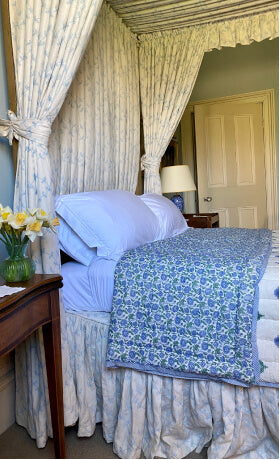 Blue patterned bespoke quilts on a bed next to antique table with daffodils