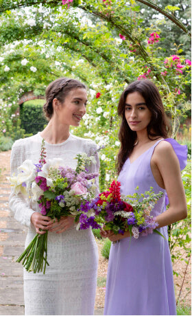 Bride and bridesmaid in lilac dress holding bouquets of flowers against garden background